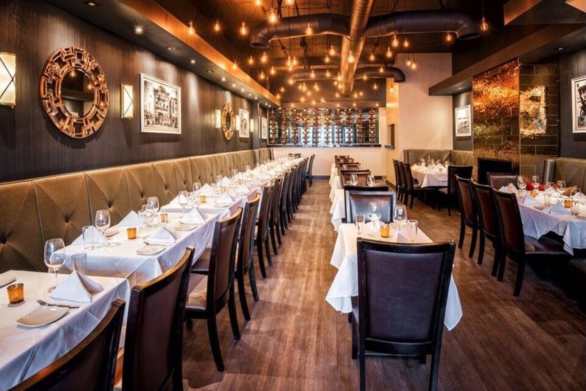 The rebuilt dining room at Greensleeves Steakhouse: a long tufted banquette down the left wall under framed photographs and round gilt mirrors, white tablecloths set for service, exposed ducts and bare bulbs overhead, and a lit wine wall at the back, much as the design set in Attachment E drew it. Source: a guest's photo on the restaurant's public Yelp listing.
